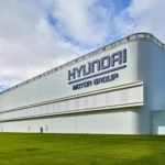 a-corner-of-an-enormous-low-white-building-is-visible-against-a-blue-sky-with-white-swirling-clouds-and-green-grass-in-the-fore